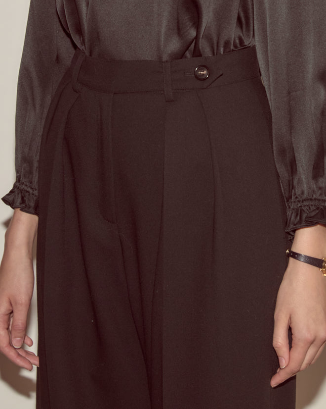A person wearing Astor Port Trousers—high-waisted, heavyweight wool pants—with a dark, long-sleeved blouse featuring ruffled cuffs.