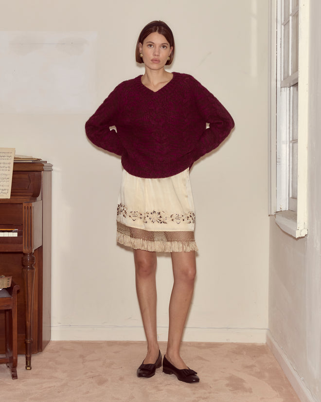 A woman stands indoors near a piano, hands on hips, wearing the ivory silk Phantasm Fringe Skirt and a maroon sweater, looking at the camera.