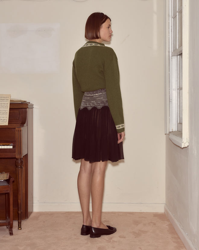 A woman in a green sweater and Ensemble Skirt stands indoors near a window, facing away from the camera.