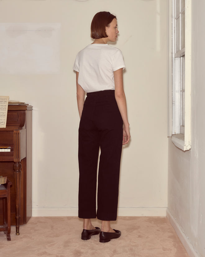 A woman wearing a white shirt and Denim Sailor Trousers - Black stands facing a corner near a window and a piano.