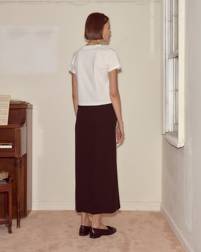 A woman indoors facing a wall, wearing the Visage Tee and a black skirt near a piano and window, brings modernist art vibes to her surroundings.