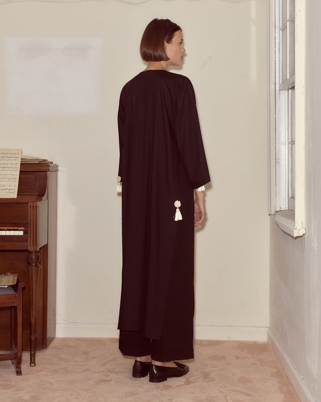 A woman in the Shadow Kaftan, a long black wool crepe dress, stands indoors by a piano near a sunlit window, facing away.