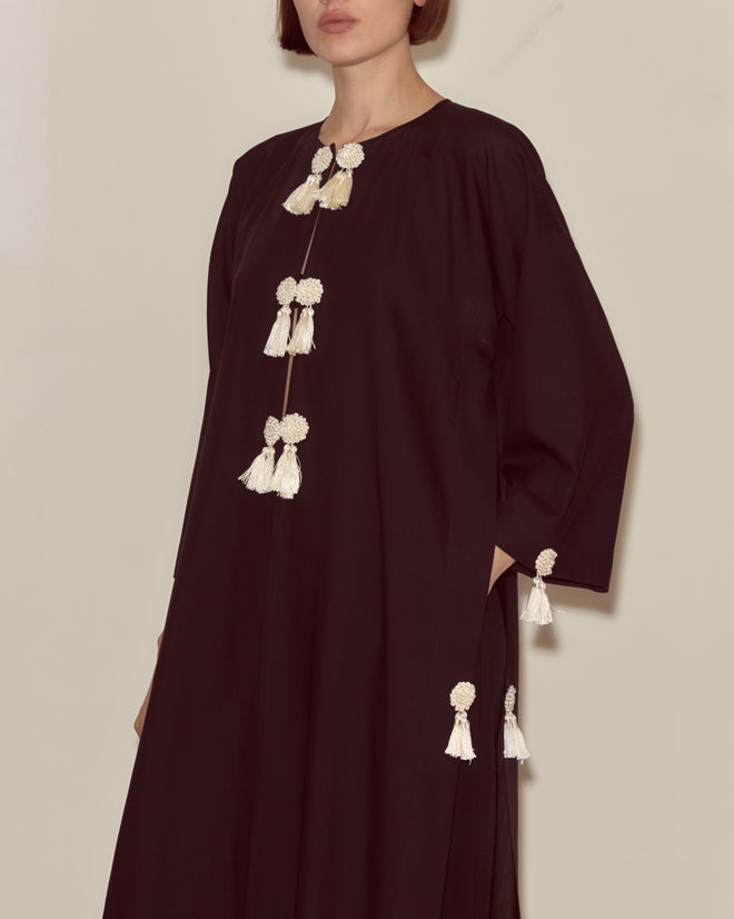 Woman wears the Shadow Kaftan, a loose, long-sleeved black dress featuring white tassel details and handy side pockets.