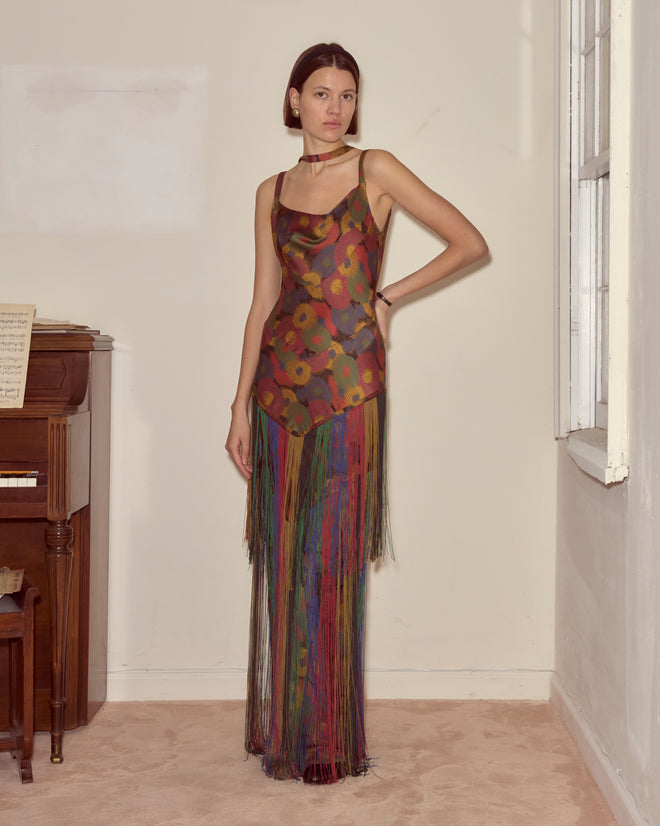 Woman in the Fruit Pit Dress stands indoors near a piano and window, hand on hip and neutral expression.