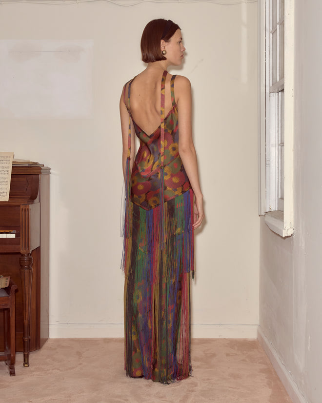 A woman wearing the Fruit Pit Dress—a colorful, backless fringe style—stands by a window in a room with a piano.