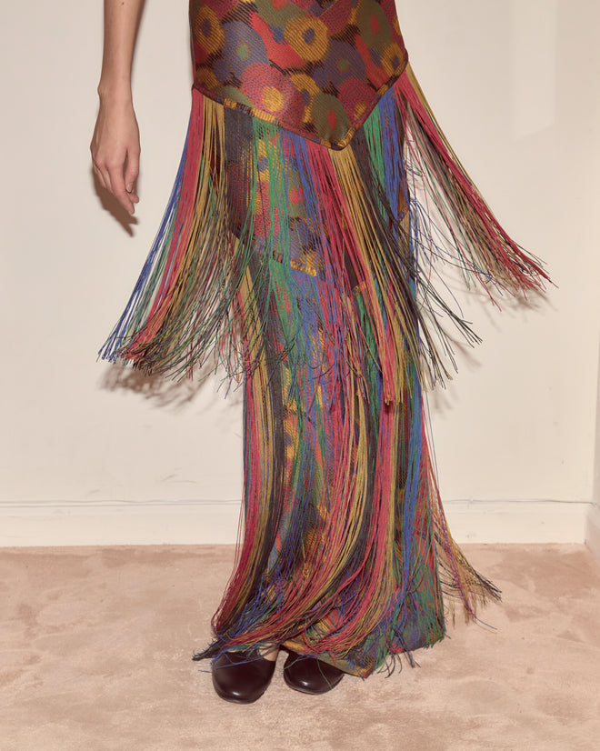 Someone stands on a beige carpet wearing the Fruit Pit Dress, a colorful fringed dress made of 100% silk with a floral pattern.