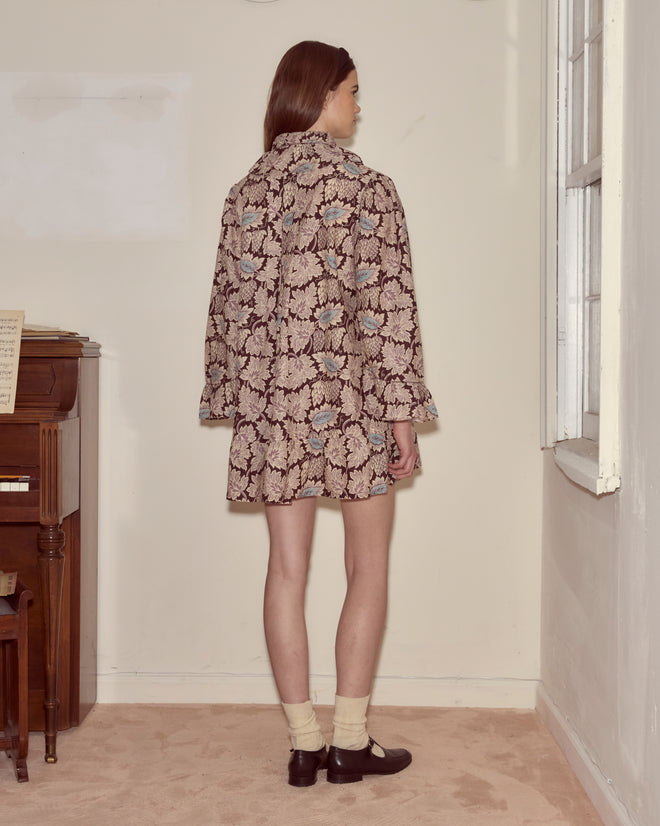 A person in a Wisteria Dress and black shoes stands facing a wall in a room with a piano and window.