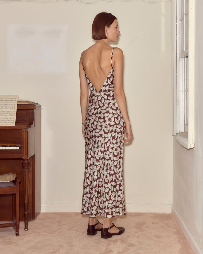 A woman wearing the Paper Birch Slip Dress, featuring a leaf-stamp pattern and low back, stands by a window in a minimal room with a piano.