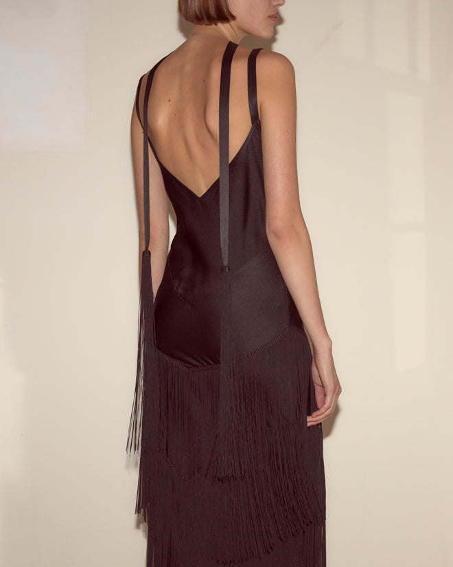A woman in the black Tango Fringe Dress stands sideways against a cream-colored wall.
