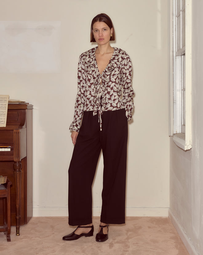 A woman wearing the Paper Birch Flounce Blouse with a leaf-stamp pattern and black pants stands in a room featuring a piano and a window.