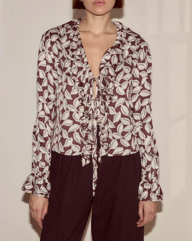 A woman wears the Paper Birch Flounce Blouse—100% silk with long sleeves, a leaf-stamp pattern, bow collar, and ruffled details—paired with black pants.