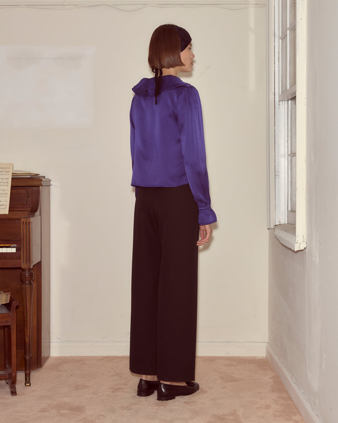 A person stands indoors, facing a wall, wearing the Dream Souvenir Flounce Blouse with a blue ribbon tie, black pants, and a black headscarf.