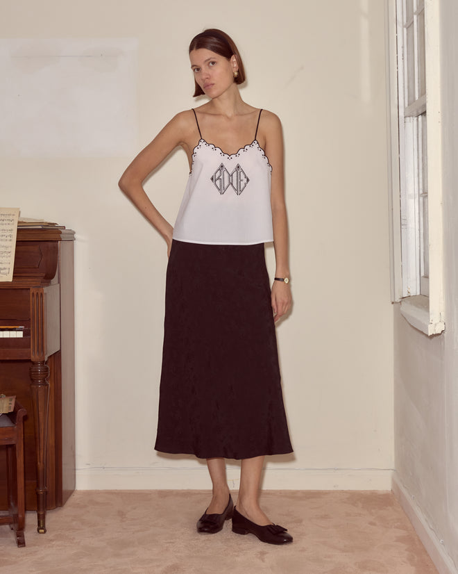 Woman in a Diamond Monogram Tank and black skirt stands indoors near a piano and window, looking at the camera.