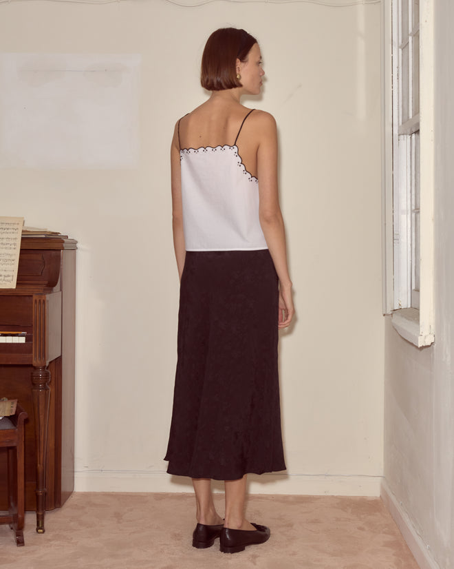 Woman standing indoors facing a wall, wearing the Diamond Monogram Tank paired with a black skirt and black shoes.