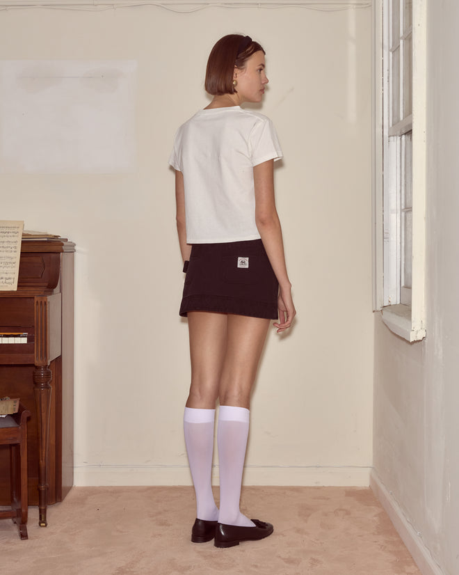A woman in a white Diamond Monogram Tee, black skirt, and knee-high socks stands by a window facing sideways.