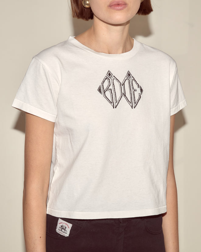 A woman facing forward wears the Diamond Monogram Tee, a white shrunken 100% cotton top with a black geometric design, paired with dark pants.