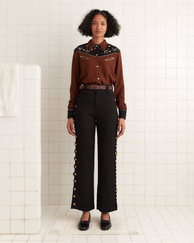 A person in a brown blouse and black Abilene Trousers adorned with studded felt flowers stands gracefully in a tiled room.