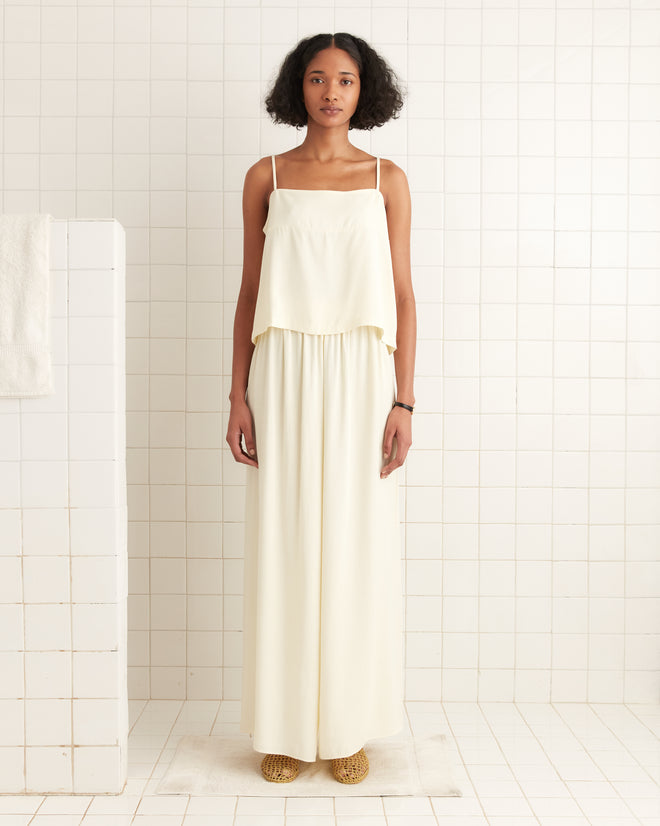 Woman wears Roy Pants in white, a loose, airy style inspired by 1920s beach pajamas, standing in a tiled white room facing the camera.