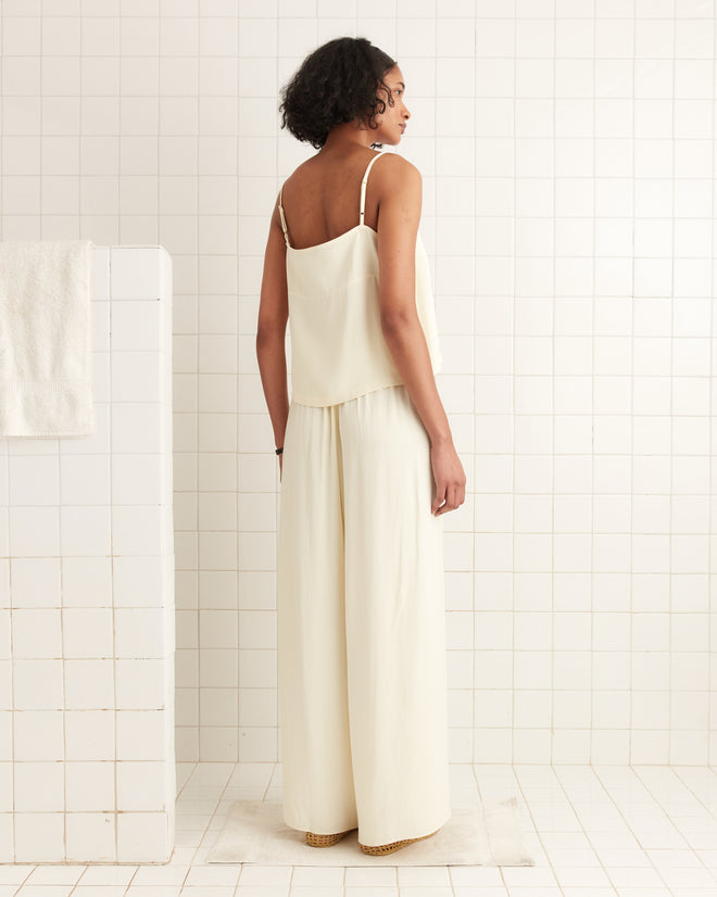A woman stands in a white tiled room wearing wide-leg Roy Pants in White and a loose, cream-colored sleeveless top, channeling the relaxed style of 1920s beach pajamas.