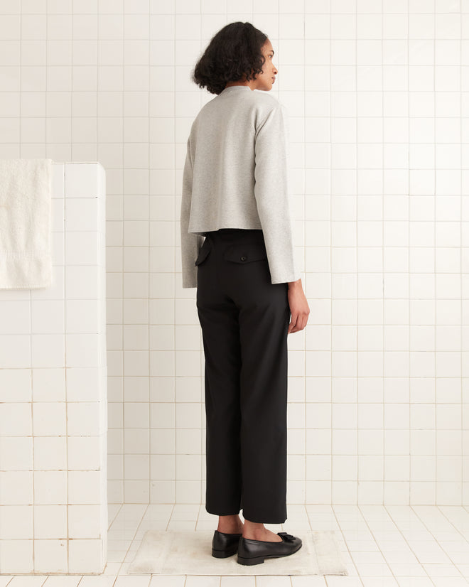 A person stands in a tiled room wearing a Heathered Cardigan - Grey, black pants, and black shoes, facing away from the camera.