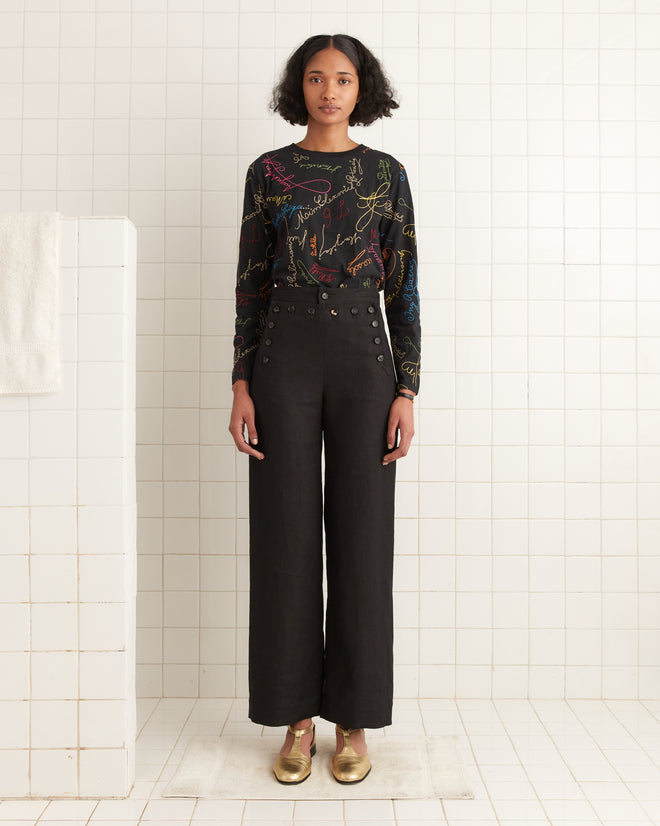 A person stands in a tiled room, wearing a Signature Scrawl Long Sleeve Tee, high-waisted black pants, and gold shoes. Their outfit is the fashion equivalent of an autograph blanket—unique and eye-catching, with each piece adding a distinct signature style.