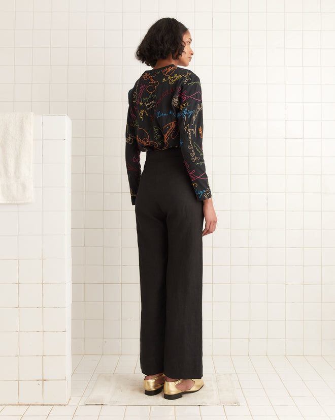 A person wearing the Signature Scrawl Long Sleeve Tee, featuring a black base with colorful patterns, stands in a tiled room, looking away. Made from 100% cotton.
