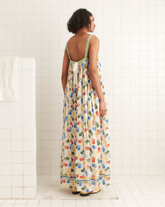 A person in a sleeveless Flower Stamp Dress, exuding vintage charm with its floral pattern, stands in a white tiled room, facing away from a white wall on the left.
