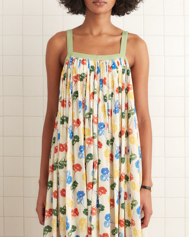 A person wearing the Flower Stamp Dress, featuring a colorful floral pattern with green straps and made from 100% cotton, stands against a tiled wall.