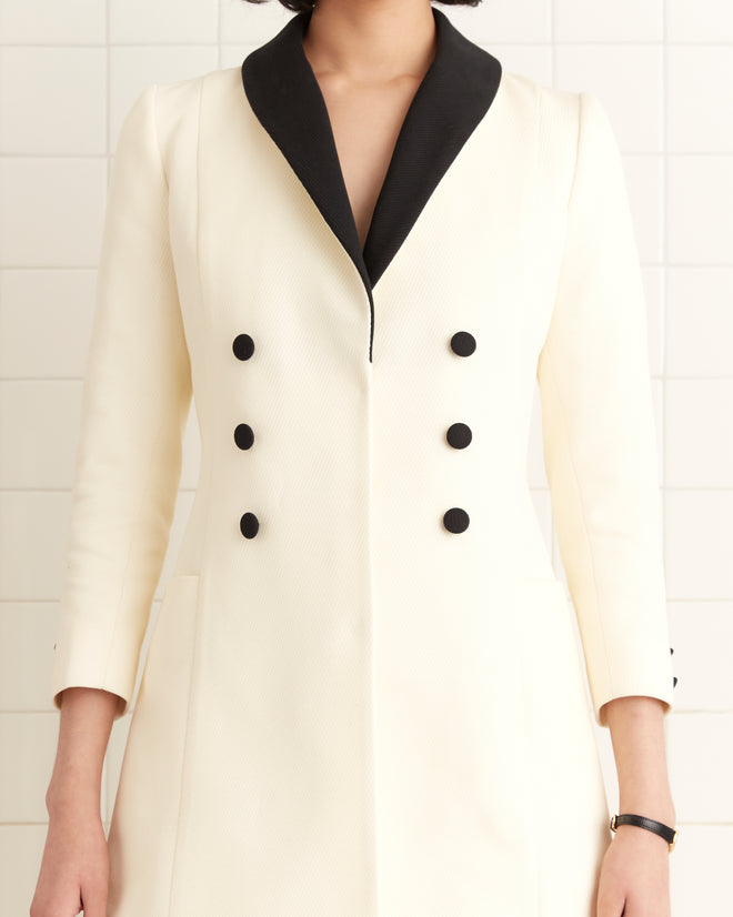 Woman wearing the Seymour Dress, an ivory wool twill jacket dress with a cream double-breasted design, black buttons, and a black collar.