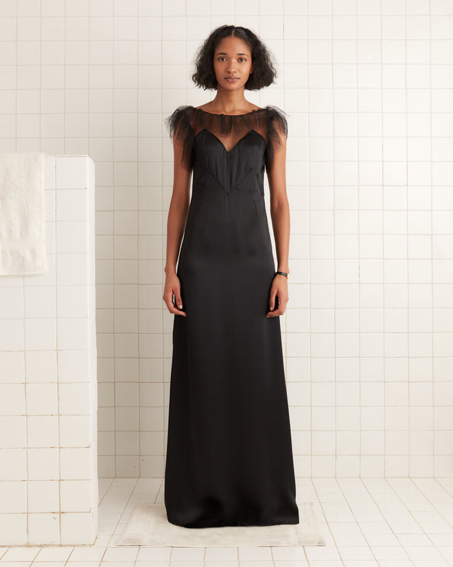 A woman wearing the Prima Dress, a long black evening gown with an illusion neckline and sheer tulle detail, stands in a white-tiled room.