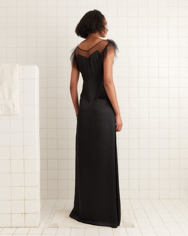 A woman wearing the Prima Dress, a long black silk gown with an illusion neckline, stands with her back to the camera in a white tiled room.