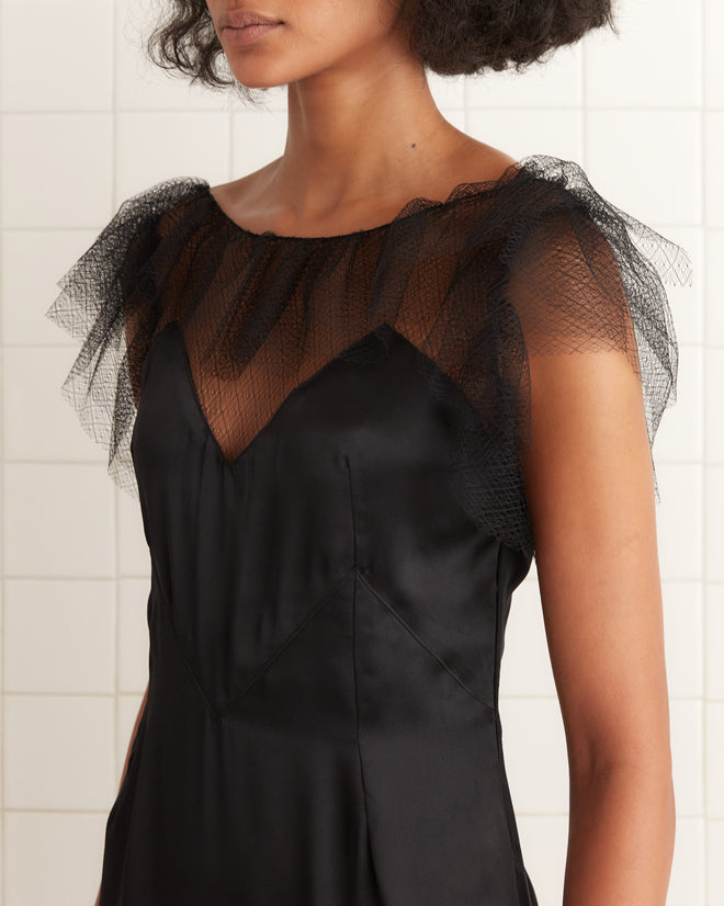 Woman wearing the Prima Dress—a black silk gown with sheer tulle ruffle and illusion neckline—stands against a tiled background.