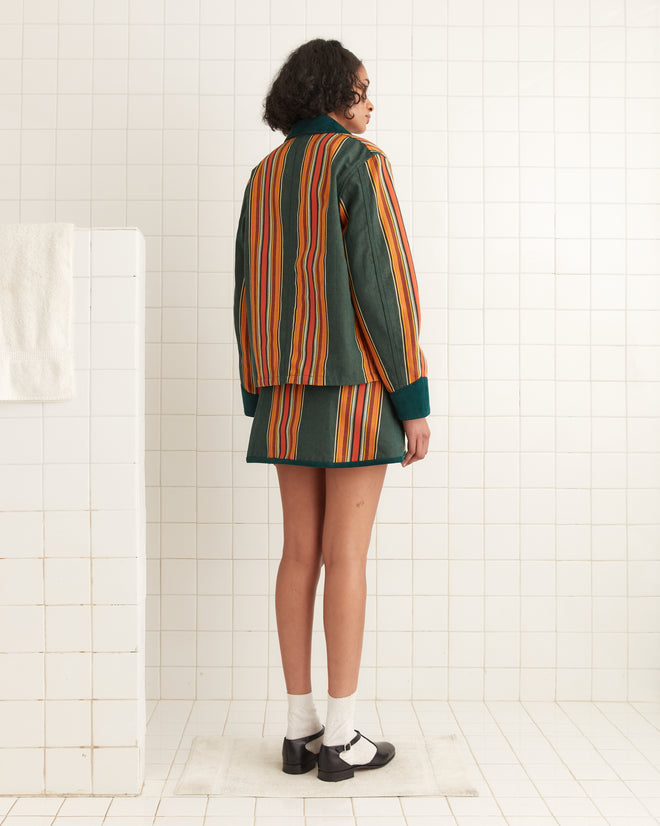 In a tiled room with a bar draped in a towel, someone wears the Parakeet Stripe Craft Jacket and matching skirt from a classic workwear ensemble. Crafted from 100% cotton, the jacket blends style and comfort effortlessly.