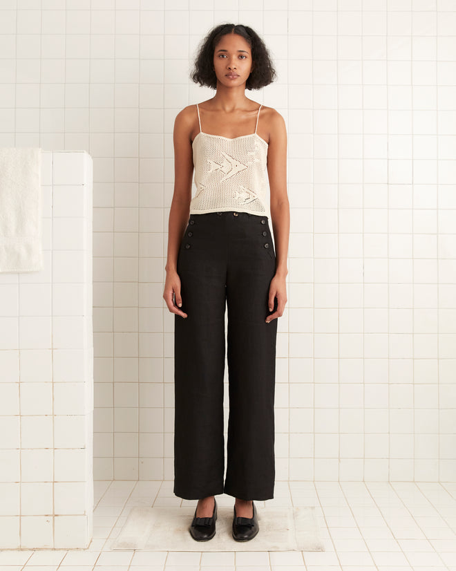 A woman wearing the Angel Fish Tank - Cream, a knit lace tank top with an angel fish pattern, pairs it with black wide-leg pants while standing in a white tiled room.