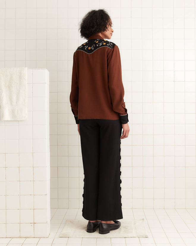 A person wearing the Abilene Western Shirt, featuring a brown hue with floral accents and pearl snap buttons, is paired with black pants. They stand on white tiles, evoking a scene from a 1960s Western TV show, facing away.