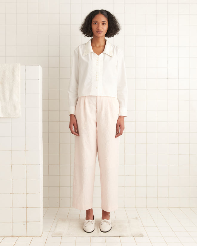 A person in a Crosshatch Shirt with antique pearl buttons and light pink pants stands on a rug in a tiled room near a wall with a towel.