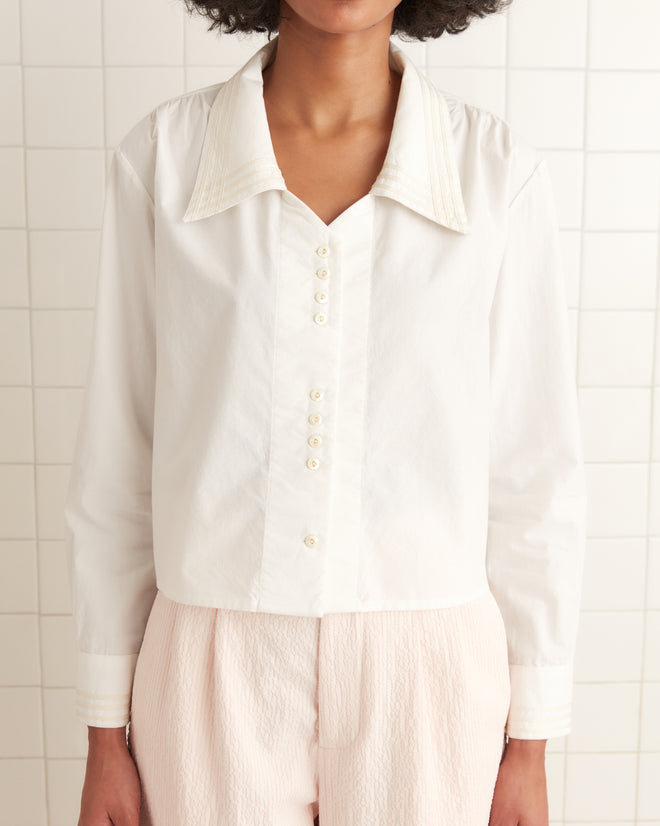 Against a tiled wall stands a person wearing light pants and the Crosshatch Shirt, a sailor-style top featuring pristine white fabric and antique pearl buttons. Face not visible.