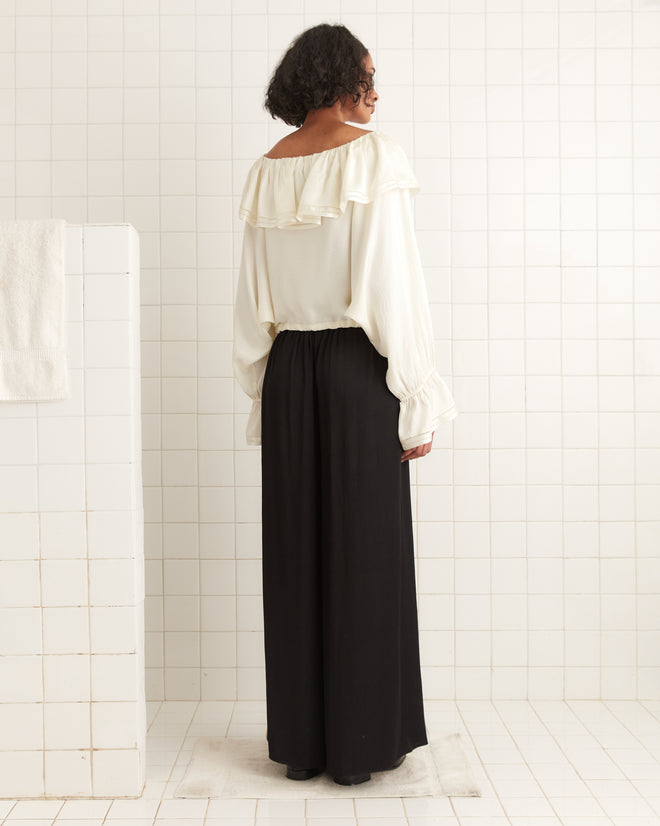 A person stands in a white-tiled room, wearing a Satin Claret Blouse - White made of 100% silk, featuring an elegant ruffled collar, paired with a long black skirt as they face away from the camera.