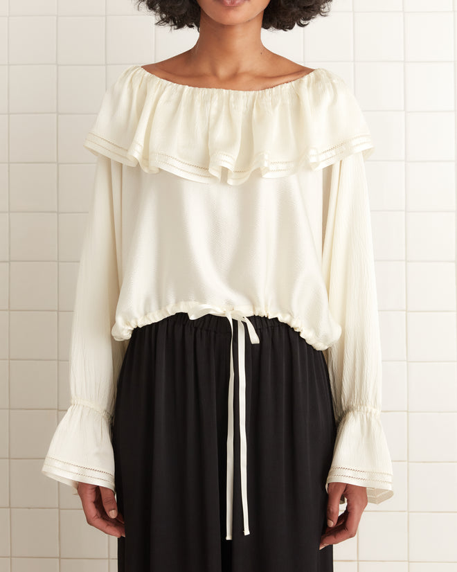 A woman in a Satin Claret Blouse - White with an elegant ruffled collar and an off-the-shoulder design, paired with a black skirt, stands against a tiled wall.