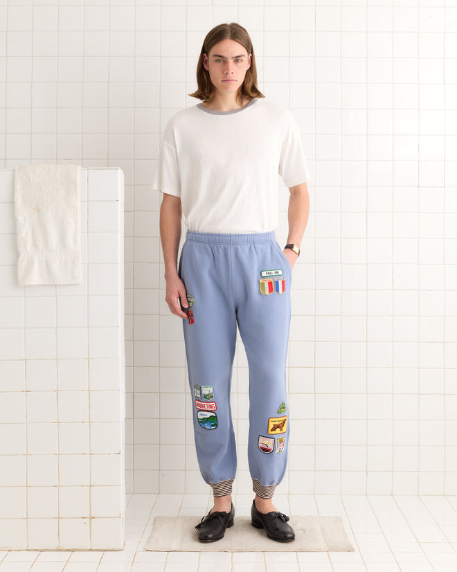 A person in a white shirt and Warmup Patch Joggers stands on a white tiled floor, with the joggers' vintage patches adding a unique touch to the scene.