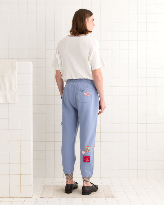 A person stands in a tiled room wearing a white shirt and Warmup Patch Joggers, facing away.