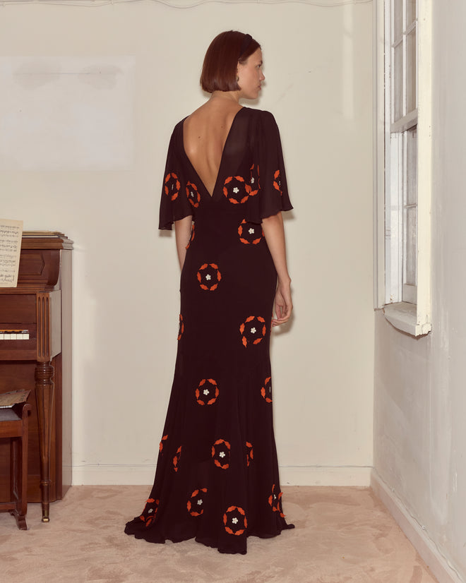 A woman stands by a window with her back to the camera, near an upright piano, wearing the Zoetrope Appliqué Dress in black silk georgette with orange patterns.