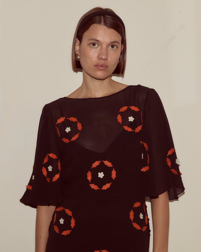 A woman wears the Zoetrope Appliqué Dress, a black silk georgette piece with orange and white floral patterns inspired by 1930s daywear, set against a plain light background.