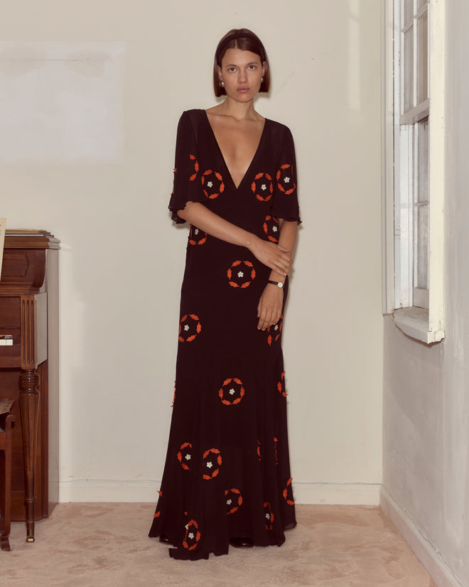 A woman stands indoors near a window and a piano, wearing the Zoetrope Appliqué Dress—a black silk georgette 1930s day dress with red and white circular patterns.