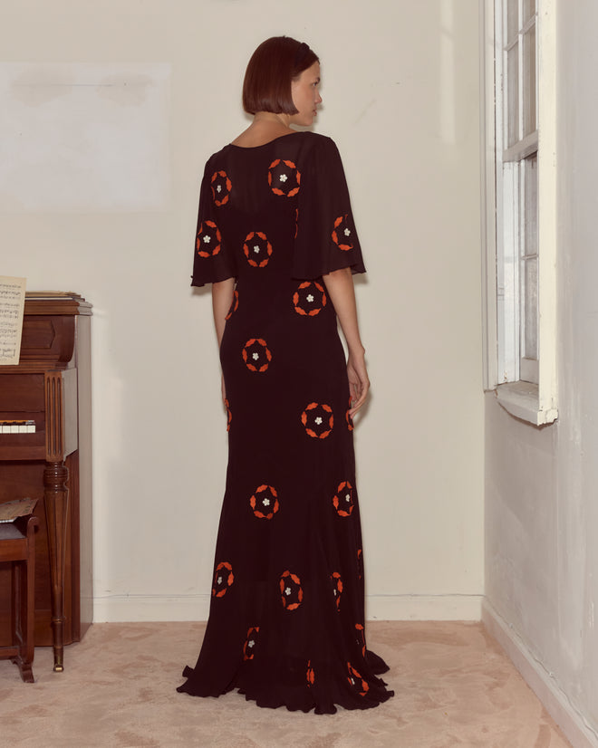 A woman wearing the Zoetrope Appliqué Dress, made from black silk georgette with red and white details, stands by a piano in a bright room, facing away.