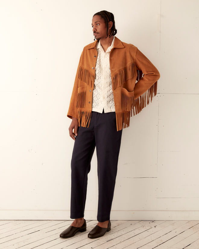Wearing the Appalachian Jacket in Camel, a white patterned shirt, navy pants, and brown shoes, this individual stands by a white wall, evoking a classic Western-inspired look.