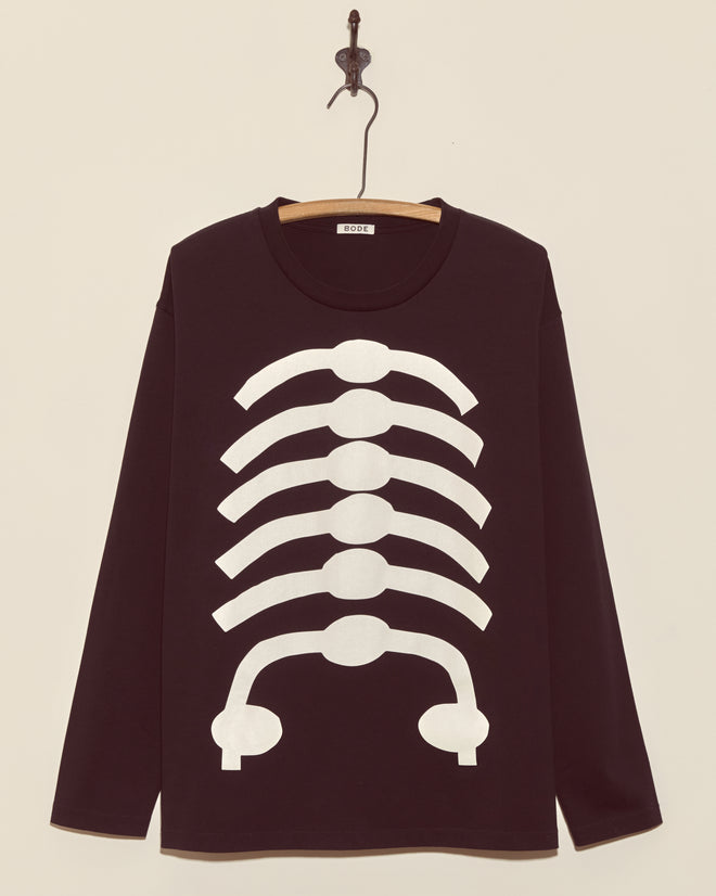 The Skeleton Long Sleeve Tee is a black shirt with a white ribcage graphic, displayed on a wooden hanger against a cream wall. This vintage tee features a boxy fit, making it perfect for your spooky season style.