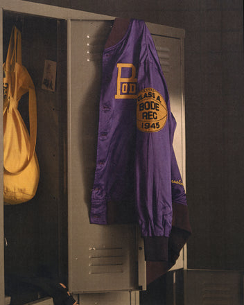 The Athlete's Rally Jacket, a vintage purple basketball championship jacket, hangs on a gray locker next to a yellow bag.