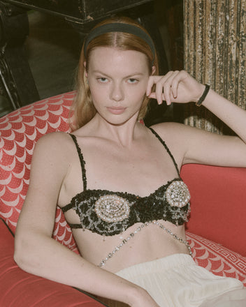 A woman with light hair sits on a red patterned chair, wearing the Oyster Bra - Black with hand beadwork and a cream skirt.