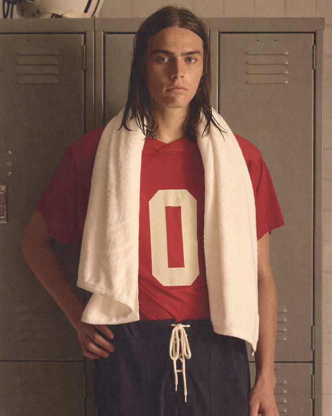 A young athlete wearing the Scrimmage Jersey - Red with a white "0" and a towel over his shoulders stands by lockers.
